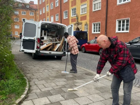 Zwei Mitarbeiter von BTS Bavaria fegen den Gehweg vor einem Münchner Wohnhaus, daneben der beladene Transporter mit zerlegten Holzmöbeln
