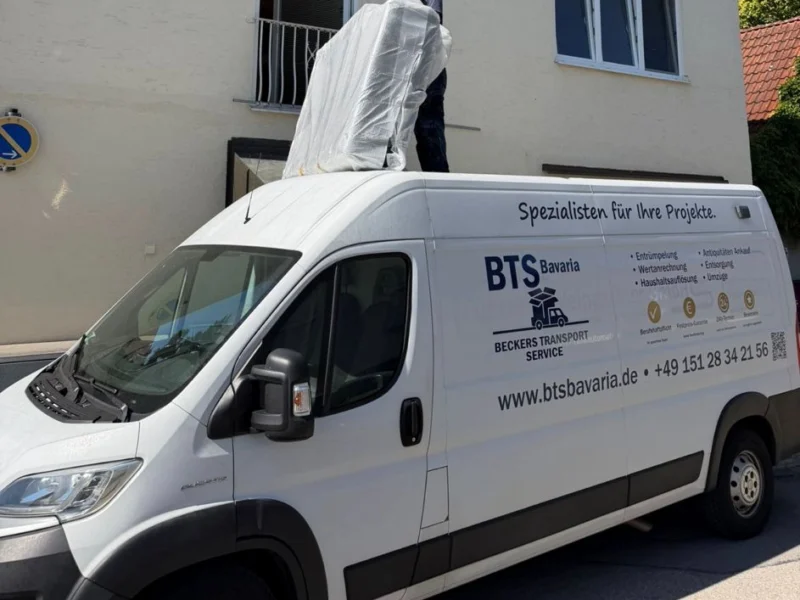 Mitarbeiter von BTS Bavaria verladen ein verpacktes Möbelstück auf dem Dach des beschrifteten Firmentransporters