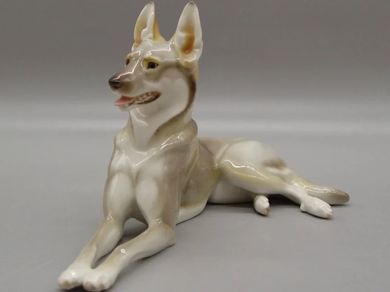 Porzellanfigur eines liegenden Schäferhundes vor neutralem Hintergrund